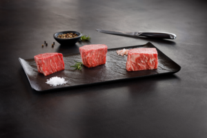 A sleek black rectangular plate displays three cuts of richly marbled Wagyu beef, showcasing the distinct marbling and high quality of the meat. The plate is garnished with sprigs of fresh rosemary, coarse salt, and a small dish of peppercorns, while a sharp knife rests in the background on a dark, elegant surface. The presentation emphasizes the luxurious and premium nature of the Wagyu beef.